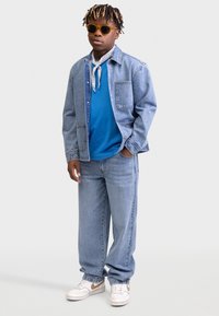 Chaqueta de mezclilla, camiseta azul, jeans de mezclilla claros y zapatillas blancas con acentos en beige. Accesorios con gafas de sol amarillas y un pañuelo blanco.