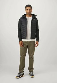 Mand iført sort hættejakke i puffer-stil over hvid skjorte, olivengrønne cargobukser og sorte, hvide og beige sneakers, stående imod en ensfarvet baggrund.