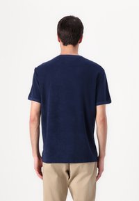 Mann in einem dunkelblauen kurzärmeligen Hemd und beigen Hosen, der mit dem Rücken zu einem schlichten weißen Hintergrund steht.