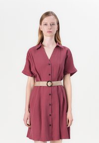 Robe boutonnée bourgogne à manches courtes et décolleté en V. Elle présente une ceinture tissée avec une boucle ronde, un tissu lisse et une silhouette fluide.