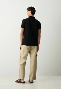 Camiseta polo negra, pantalones cargo beige y sandalias marrones. La camiseta tiene mangas cortas, mientras que los pantalones cuentan con bolsillos laterales y un corte relajado.