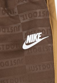 Rjavi atletski šortai z teksturirano tkanino, ki vsebuje ponavljajoči se vzorec "JUST DO IT". Bel Nike logotip na levi strani blizu pasu.