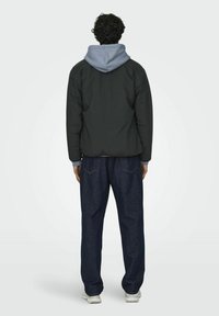 Black padded jacket with a grey hood, worn over a light blue shirt, paired with loose dark denim jeans and white sneakers.