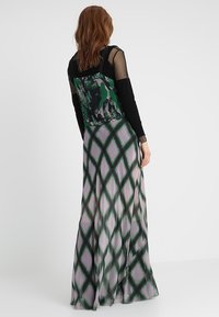 Une femme aux cheveux auburn porte un haut noir à manches longues avec des manches transparentes et une robe sans manches à motifs verts avec une longue jupe à imprimé diamant.