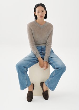 Nuori nainen, jolla on lyhyet mustat hiukset, istuu valkoisella laatikolla. Hänellä on päällään beige pörröinen neule, siniset farkut ja tummanruskeat loafersit valkoista taustaa vasten.