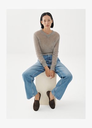 Nuori nainen, jolla on lyhyet mustat hiukset, istuu valkoisella laatikolla. Hänellä on päällään beige pörröinen neule, siniset farkut ja tummanruskeat loafersit valkoista taustaa vasten.