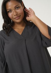 Blouse gris foncé avec un décolleté en V, motif à pois texturé et manches 3/4 amples. Le tissu semble doux, léger et tombe doucement.