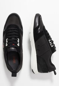 Ett par svart- och vitfärgade Hugo Boss-sneakers med snörningsdesign, paneler i mocka och läder samt en kraftig vit sula på vit bakgrund.
