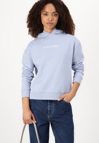 Svijetloplavi dukserica od glatkog materijala, sa kenguruh džepom, rebrastim manžetama i bijelim logom na prsima. Nosi se s tamnoplavim trapericama.