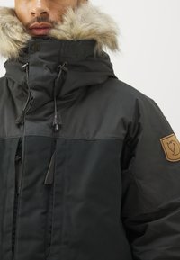 Sort isoleret jakke med en stor faux fur hætte, der har et struktureret yderlag og et læderpatch på ærmet.