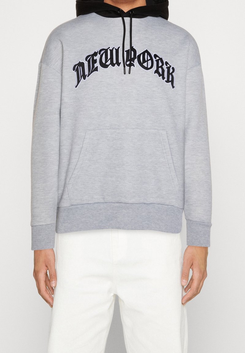 Personne portant un sweat à capuche gris clair avec le texte noir "NEW YORK" sur la poitrine et un pantalon blanc, debout devant un fond uni.