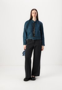Chaqueta bomber de cuero azul verdoso con cierre de cremallera, cierre a presión y diseño corto, combinada con pantalones negros de pierna ancha y un bolso azul pequeño.