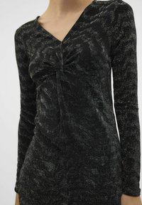 Vestido negro de manga larga con escote en V y detalle de frente retorcido. El material tiene un acabado texturizado y brillante. Silueta recta.
