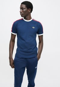 Vyras dėvi mėlyną trumpomis rankovėmis dėvimą Adidas marškinėlį ir derančias kelnes, žiūrintis į kairę su rankomis iš dalies kišenėse priešais vienspalvį foną.