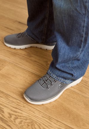 Harmaat urheilushoesit, joissa on verkkopäällinen, valkoinen pohja ja joustavat nauhat. Yhdistetty sinisiin farkkuihin puulattialla.