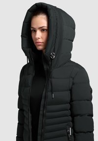 Padded čierna bunda s vysokým golierom a prehnanou kapucňou. Obsahuje predný zips, detail s šnúrkami a nášivku s logom na rukáve.