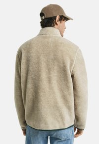 Beige fleece jas met een hoge kraag, zachte textuur en een iets oversized fit, gecombineerd met lichtblauwe denim jeans.
