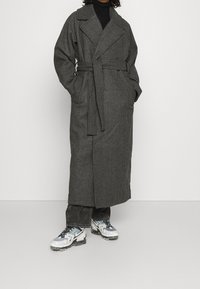 Lang grijs geblokt jasje met een riem in de taille, brede revers en zijzakken, gecombineerd met donkere denim en contrasterende sneakers.