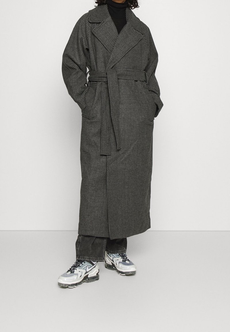 Lang grijs geblokt jasje met een riem in de taille, brede revers en zijzakken, gecombineerd met donkere denim en contrasterende sneakers.