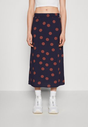 Jupe midi bleu marine avec de grands pois couleur rouille. Dotée d'une taille élastique, elle descend jusqu'au mollet, assortie à des baskets blanches.