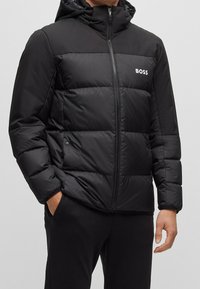 Svart puffjacka med dragkedja fram, huva och logotypdetaljer. Har quiltade sektioner och ett mjukt, glänsande tyg. Bekväm passform.