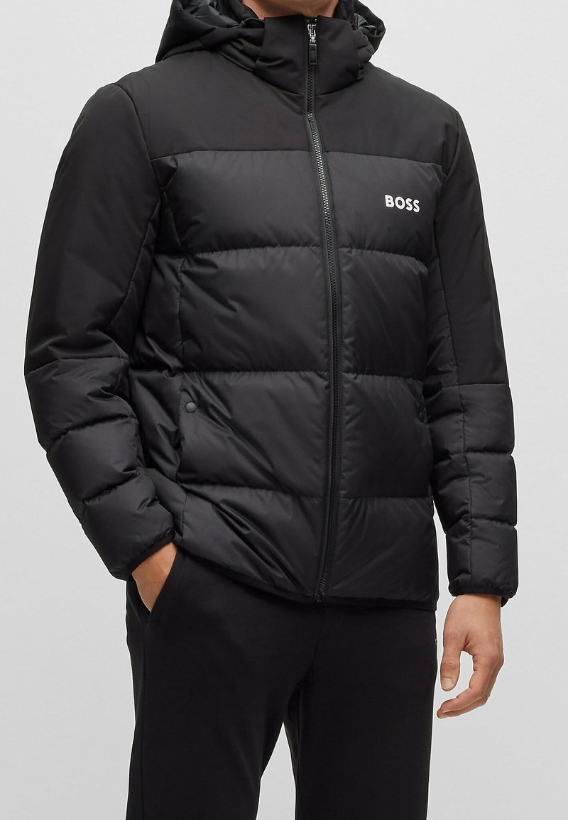 Svart puffjacka med dragkedja fram, huva och logotypdetaljer. Har quiltade sektioner och ett mjukt, glänsande tyg. Bekväm passform.