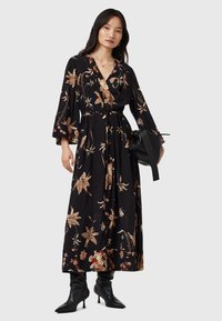 Vestido negro con estampado floral, de corte cruzado y mangas largas acampanadas, que presenta patrones de flores en beige y naranja. Combinado con un bolso negro y botas de tacón.