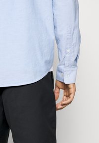 Rechter Arm und Hand einer Person, die ein hellblaues Langarmhemd und schwarze Hose trägt, vor einem schlichten Hintergrund stehend.