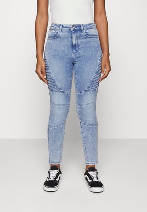 Jeans de ganga azul claro com uma cintura alta, detalhes canelados nas coxas e pernas ajustadas e afuniladas, combinados com sapatilhas pretas.