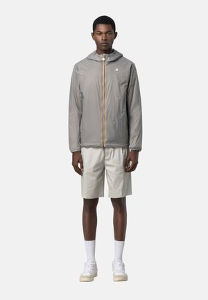 Uomo in piedi rivolto in avanti che indossa una giacca a cappuccio grigio chiaro con cerniera gialla, pantaloncini beige, calzini bianchi e scarpe da ginnastica bianche.