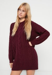 Uitgebreide wijnrode gebreide sweaterjurk met een gestructureerd kabelpatroon, geribde manchetten en een brede halslijn. Zacht, dik materiaal.