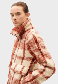 Fleece jacket with a high collar, featuring a checkered pattern in orange and cream. Zipper front and two patch pockets. Soft texture.