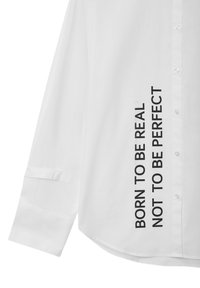 Chemise blanche à boutons avec un ourlet courbé, manches longues et texte noir sur le côté : "NÉ POUR ÊTRE RÉEL, PAS POUR ÊTRE PARFAIT."
