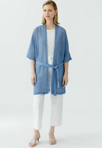 Blau gestrickte kimonoartige Strickjacke mit Bindegürtel, offener Front, kurzen Ärmeln und strukturiertem Design, kombiniert mit weißen Hosen.
