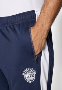 Jūras spēku sporta bikses ar baltām sānu paneļiem, ar rāvējslēdzēja kabatu un logotipu ar zvaigznēm, izgatavotas no gludas, stiepjošas auduma.