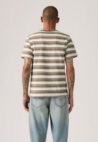 Ριγέ μπλούζα σε ελαιμί πράσινο και λευκό, βαμβακερό ύφασμα, λαιμόκοψη στρογγυλή, κοντά μανίκια. Συνδυασμένη με τζιν παντελόνι ανοιχτό μπλε. Ορατά λεπτομερή τατουάζ.