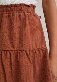 Jupe tiered couleur rouille avec un tissu texturé, dotée d'une taille froncée et de motifs floraux brodés sur le côté.