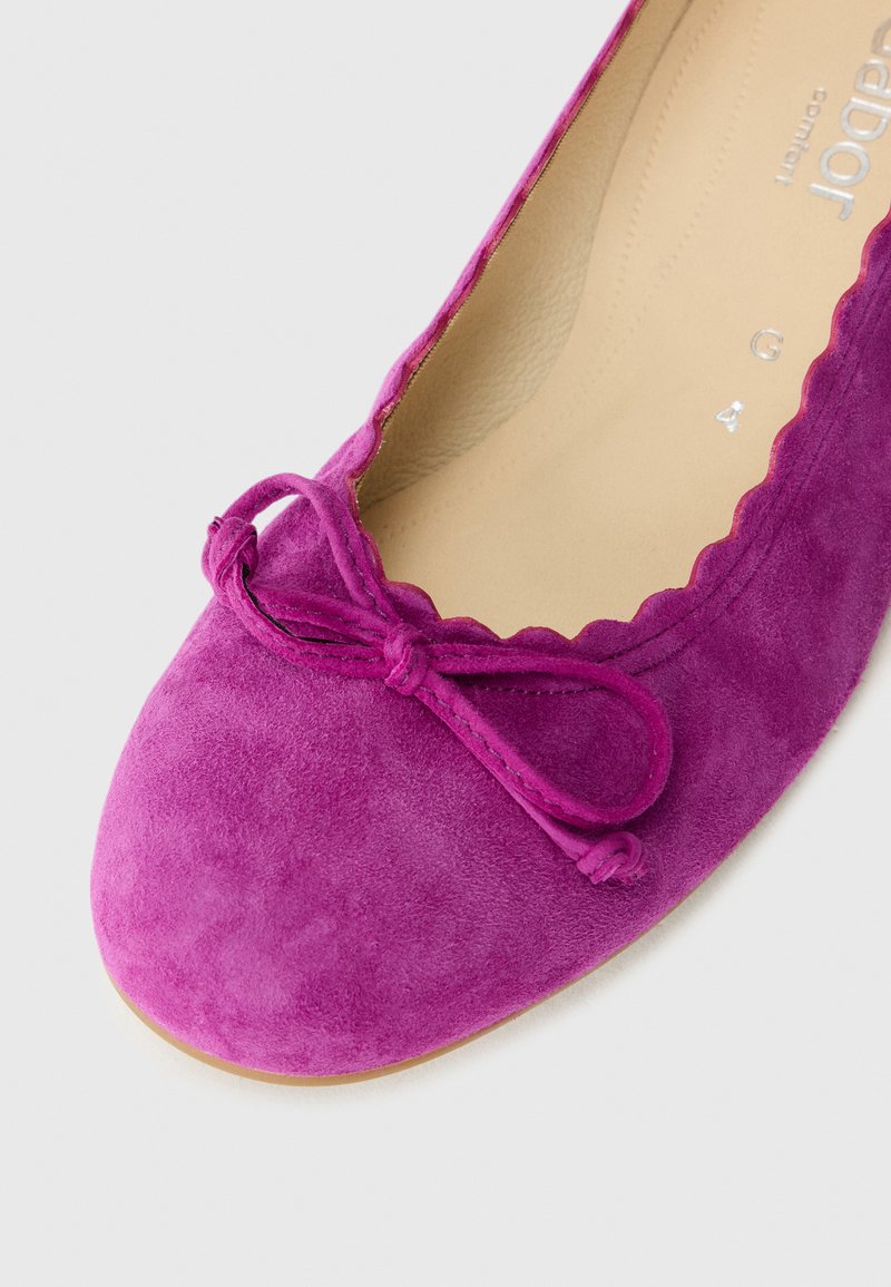 Zbliżenie jasnoróżowego zamszowego baletka z detalem kokardki na palcach, ukazujące jego falbaniasty brzeg i gładkie wykończenie wnętrza.