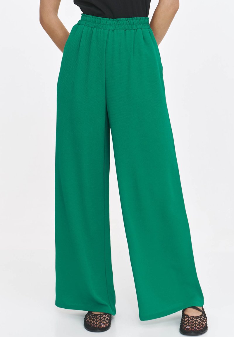 Zielone spodnie szerokie z elastycznym pasem, wykonane z lejącego materiału. Gładka tekstura, całkowita długość, zestawione z czarnym obuwiem.
