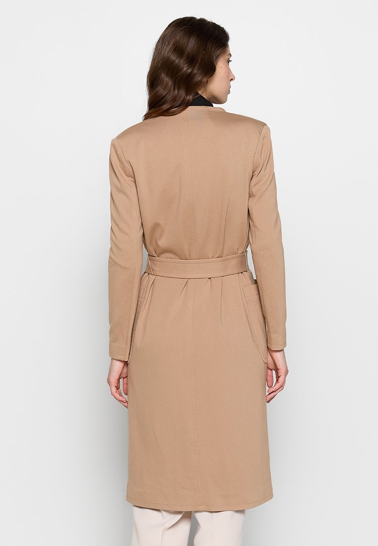 Abrigo largo beige con cintura ceñida, corte ajustado y mangas largas. Confeccionado en tejido suave, presenta un color sólido y un diseño minimalista.