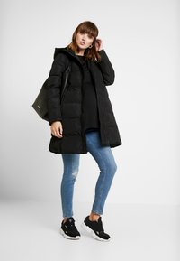 Casaco puffer preto com capuz, fecho de correr na frente, textura acolchoada, combinado com jeans skinny azuis e ténis pretos. Inclui também uma bolsa tote preta.