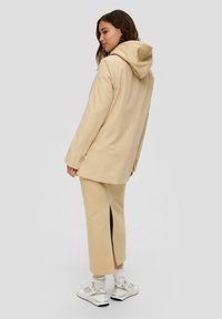 Beige pinstriped oversize takki hupparilla, yhdistettynä beigeihin halkiotaskuihin ja valkoisiin lenkkareihin. Sujuva kangas ja rento istuvuus.