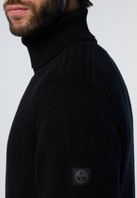 Maglione nero a collo alto lavorato a maglia con collo e dettagli sulle spalle a coste. Presenta un'etichetta con un logo circolare sulla manica sinistra.