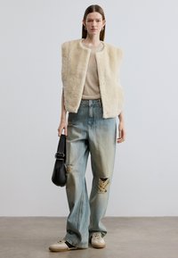 Μπεζ αμάνικο γιλέκο από συνθετική γούνα πάνω από ένα ανοιχτό καφέ τοπ, σε συνδυασμό με ανοιχτό μπλε τζιν παντελόνι με φαρδιά πόδια και φθορές, και μπεζ αθλητικά παπούτσια. Μαύρη τσάντα ώμου.