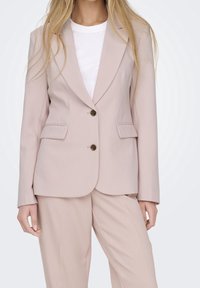 Veste rose pâle sur mesure avec un col à revers cranté, une fermeture à deux boutons et des poches avant, assortie à un pantalon sur mesure assorti.