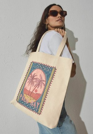 Woman wearing sunglasses and hoop earrings holds cream tote bag with colorful embroidered palm tree design and "Palm Bay" text.
