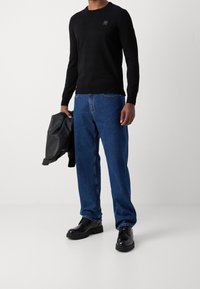 Czarny sweter z długim rękawem i okrągłym dekoltem z małym logo, połączony z niebieskimi dżinsami i czarnymi skórzanymi butami; trzyma czarną kurtkę.