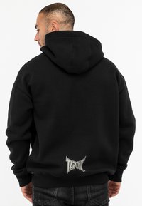 Fekete kapucnis pulóver kenguru zsebből, bordázott mandzsettával és derékpánttal. Az alsó hátrészen ezüst "TAPOUT" logóval rendelkezik.