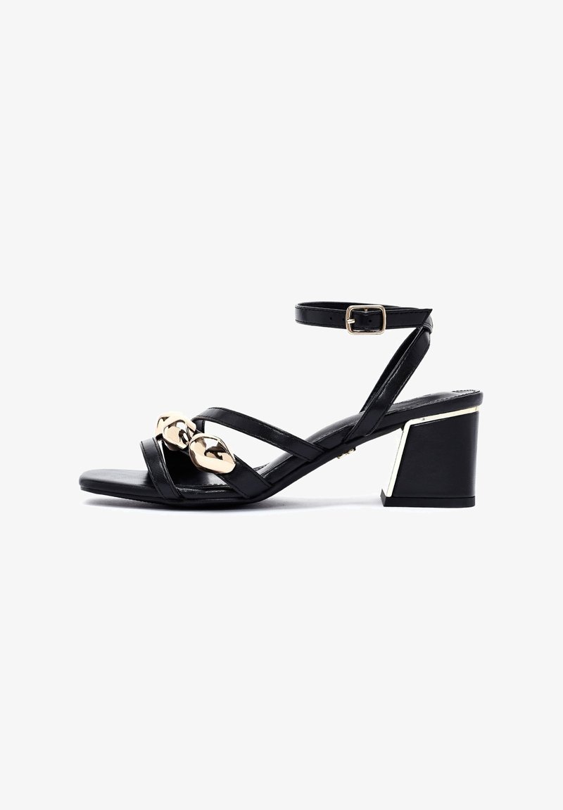 Sandalia de cuero negro con tres tiras cruzadas, acentos decorativos en dorado y un tacón grueso con un detalle metálico en el lateral.