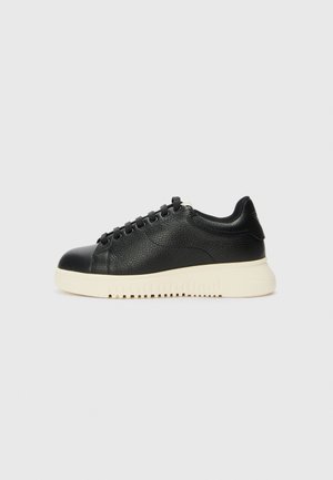 Sneaker en cuir noir avec une finition texturée, dotée d'un bout rond, de six œillets noirs et d'une semelle en caoutchouc blanche avec des rainures.